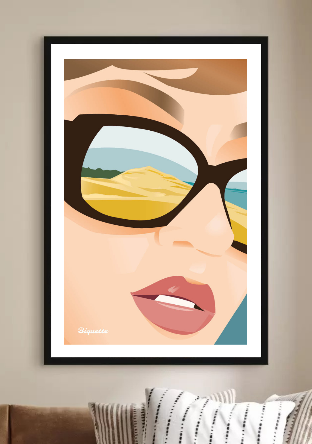 Affiche de la Dune du Pilat dans un cadre noir sobre, suspendue au centre d'un mur beige clair au-dessus d'un canapé en tissu marron glacé agrémenté de coussins à motifs géométriques noirs et blancs.