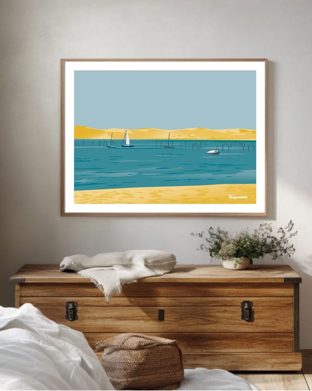 Grand cadre en bois clair affichant l'illustration de la plage du Cap Ferret et de la Dune du Pilat, suspendu au-dessus d'une commode en bois brut.