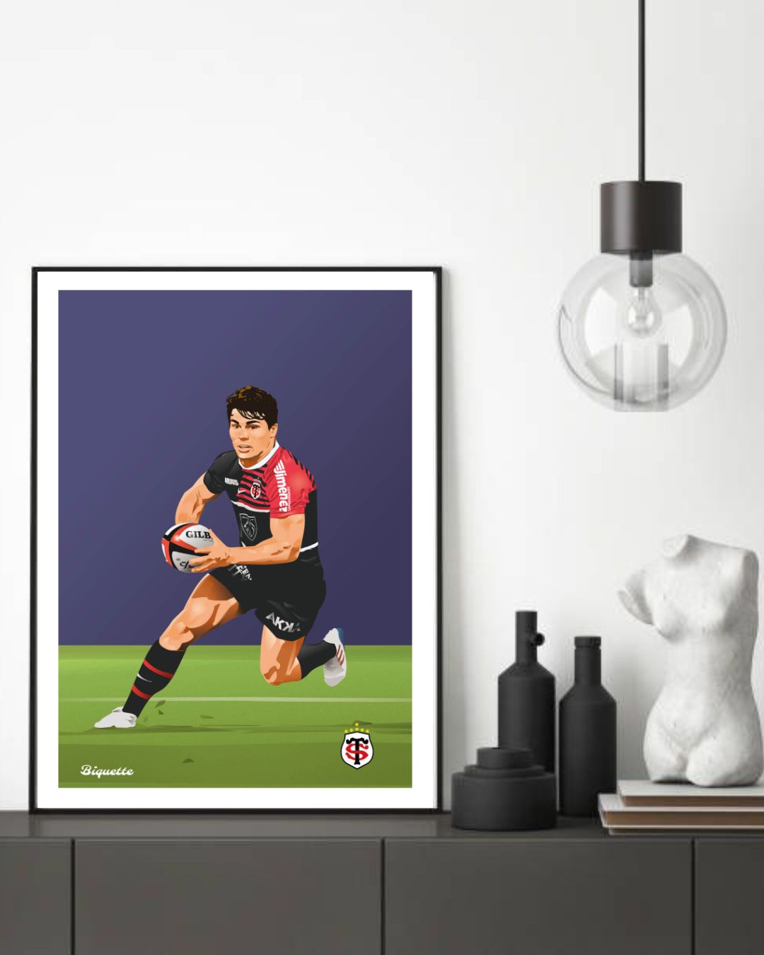 Cadre noir affichant le poster d'Antoine Dupont (Stade Toulousain) posé sur un meuble en enfilade noir mat à côté d'une petite sculpture blanche.